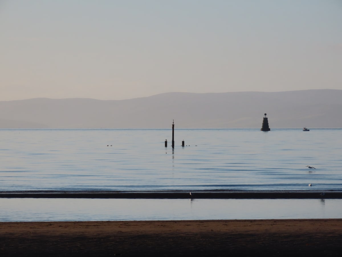 photograph from the beach on a calm day
strip of sand in the foreground
Next a strip of water follwed by another thinner strip of sand
the sea behind is limped, 3 poles and a large buoy are visiable
Bening the outline of hills
a pale sky
The whole picture is pale