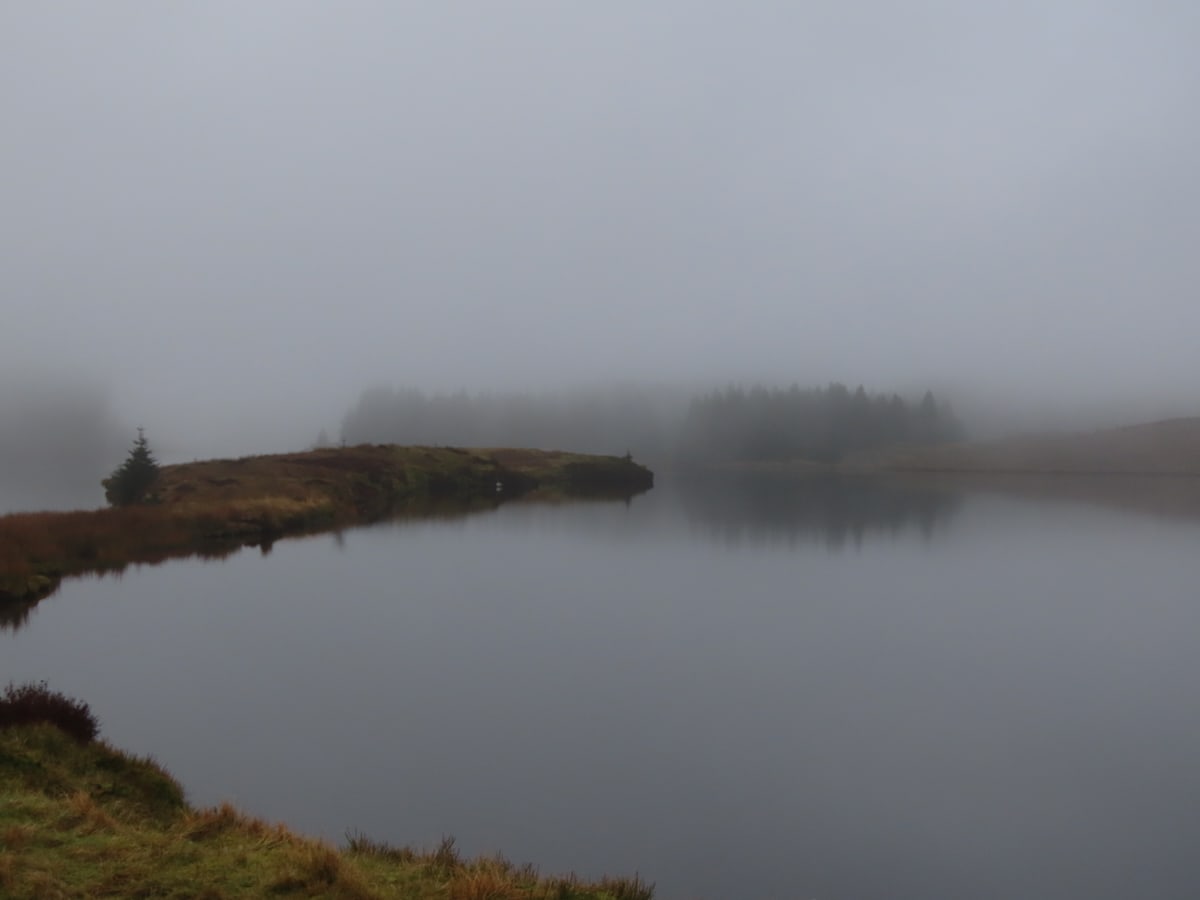 Loch Humprey in the mist. A thin peninsula arcs out from the left. Everything hazy. Conifers can be seen at the far side of the loch.