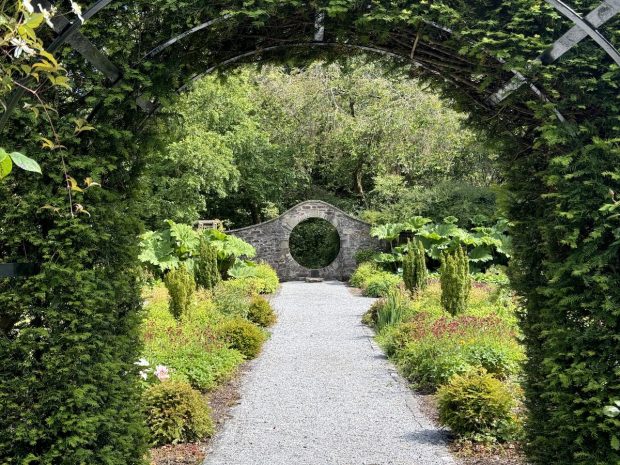 A lush garden with a gravel path, archway covered in green foliage, and a circular stone structure.
