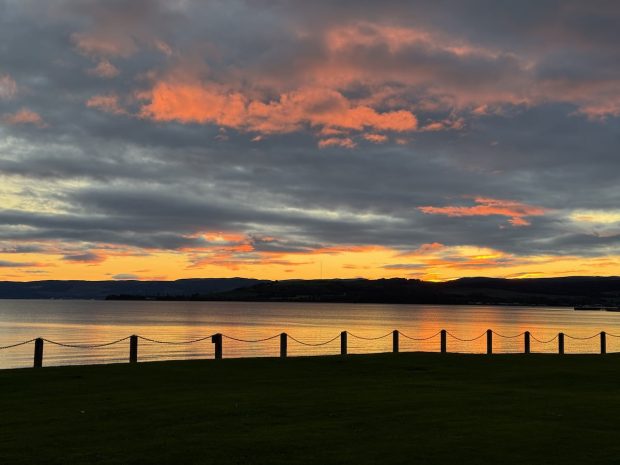 Sunset over the Clyde Estuary with dark clouds, vibrant orange and pink sky, and calm reflective water. Foreground features a grassy area with posts and chains.