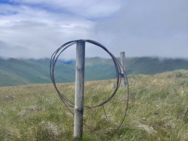 A fence post with a loop of rusty wite hanging on the top. Hills & clouds in the background.