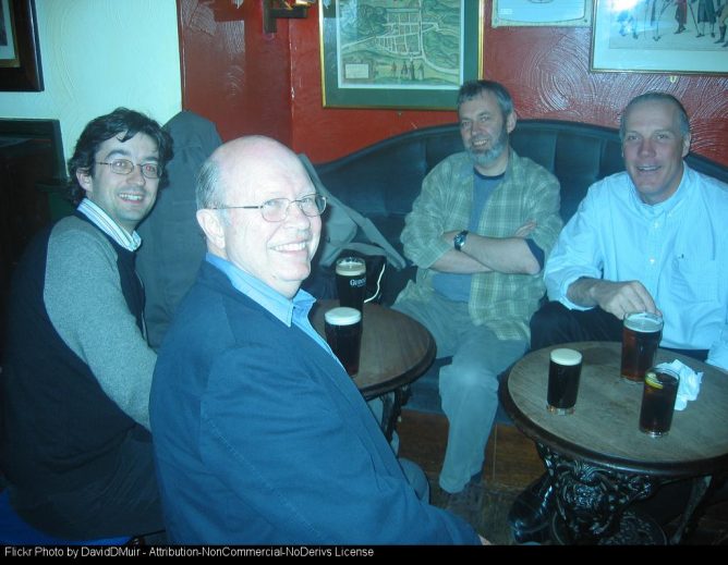 Four men sitting in a pub, smiling and talking, with pints of dark beer on two small tables. Bob, Ewan, Me & Will at the Jolly Judge.