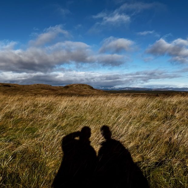 Shadows of two people cast on a field of tall grass under a blue sky with clouds.