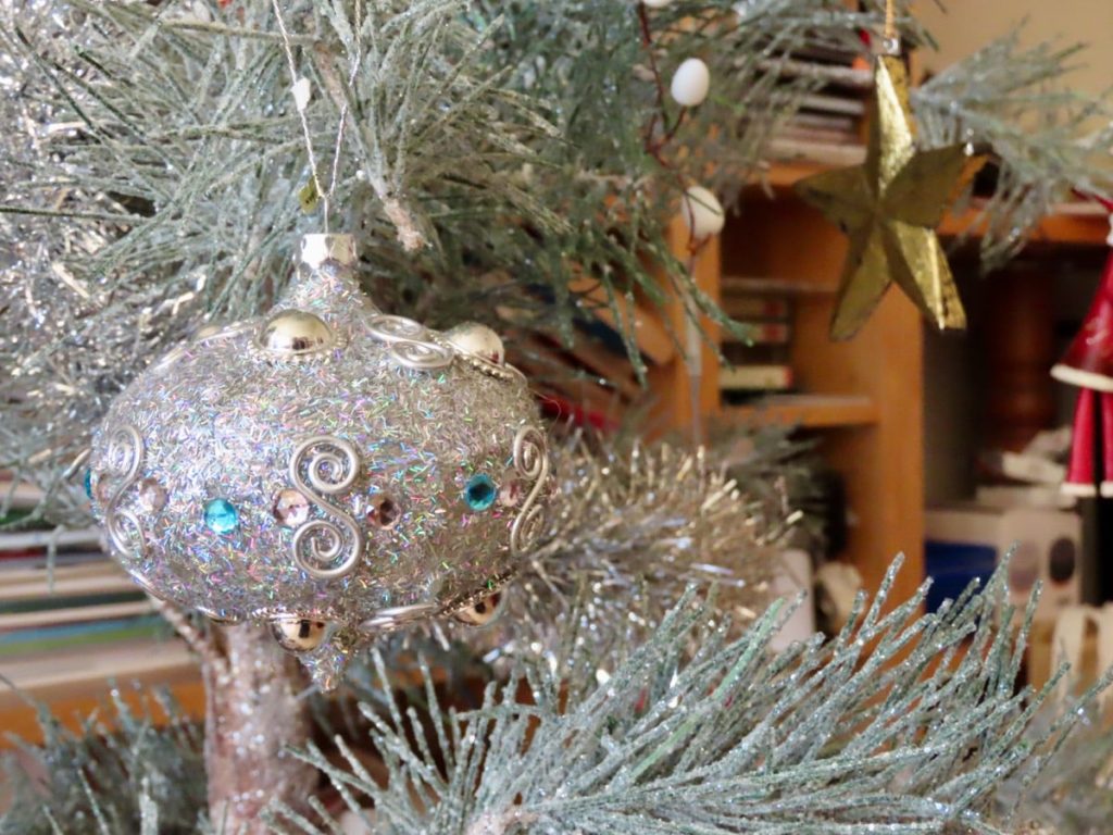 A silver spherical ornament with gems hangs on a tinsel-covered branch, with a golden star in the background.