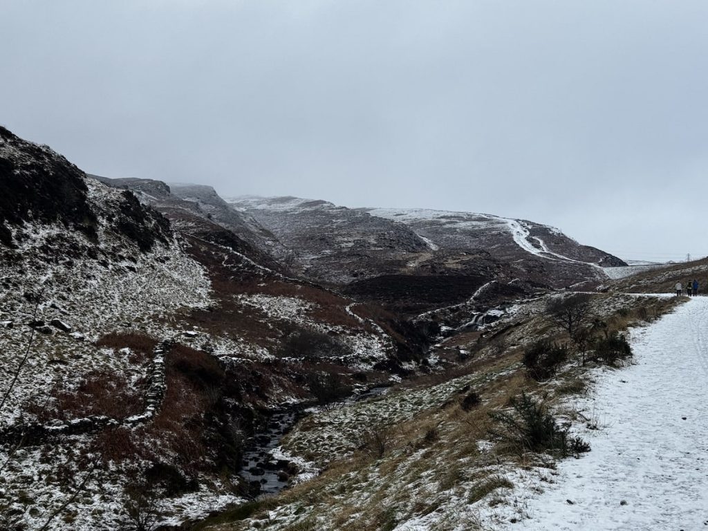 Snowy landscape with hills and a cloudy sky, featuring a path and a stream.