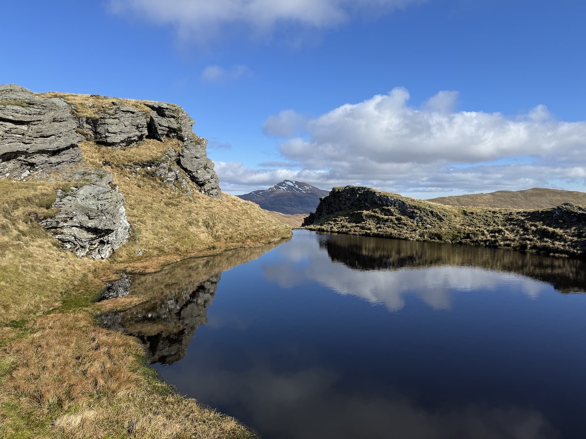 A peaceful landscape with a reflective body of water, rocky grass-covered banks, and a distant snow-capped mountain under a blue sky with white clouds.