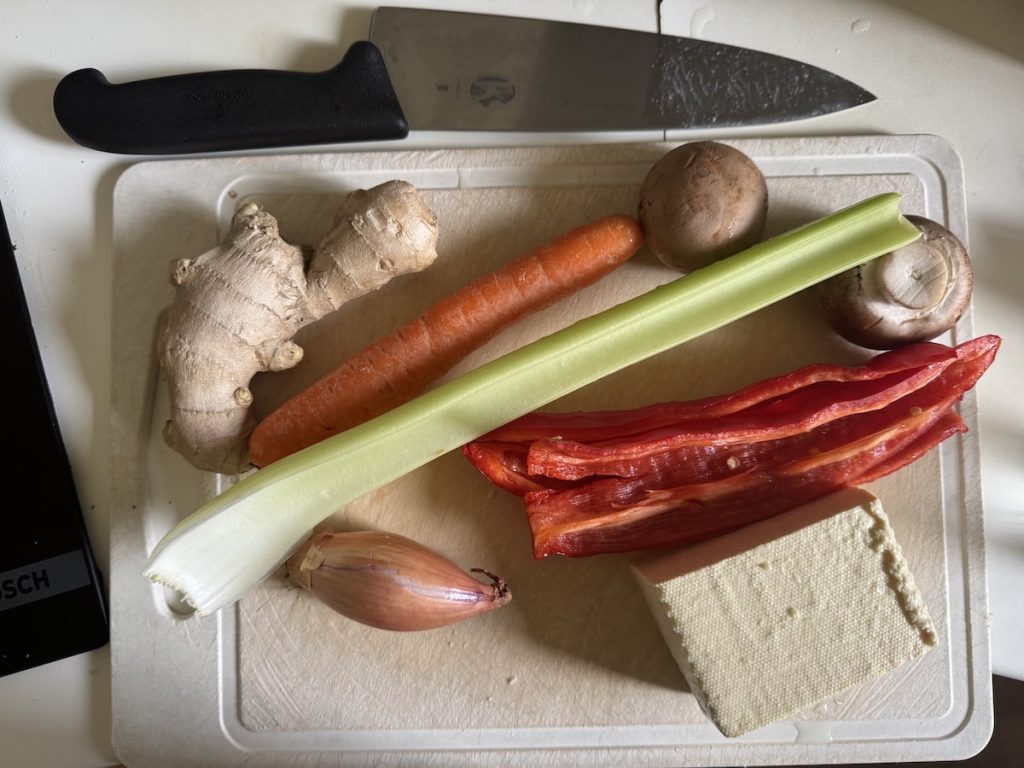 Fresh ingredients including ginger, carrot, celery, red pepper slices, mushrooms, tofu, and a shallot on a cutting board with a chef's knife.