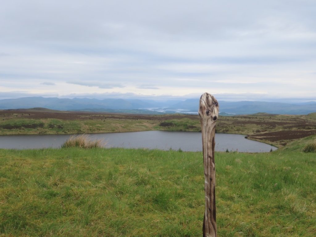A grassy hill with a wooden post, a loch, and distant mountains under a cloudy sky.