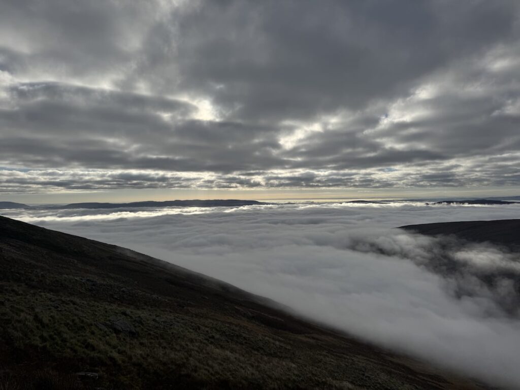 A dark mountain slope overlooks a valley filled with dense white fog beneath layered gray clouds.